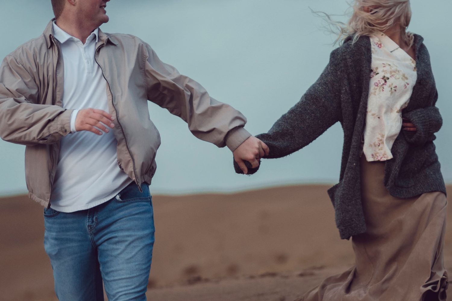 spring beach engagement session, couple run across the dunes holding hands while the wind blows their layers around.