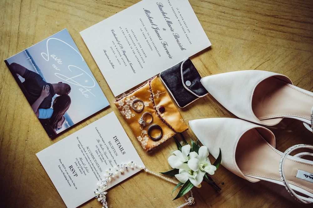 Wedding details including white bridal shoes, gold rings, invitation cards and delicate white flowers arranged on wooden floor.