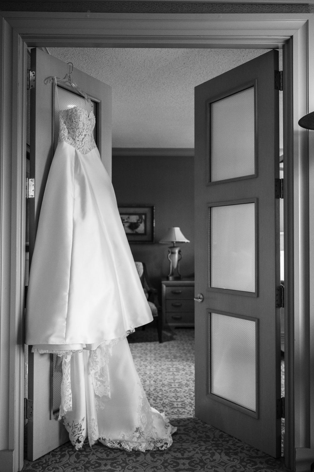 An elegant white wedding gown with lace details hangs in a doorway of a hotel room in black and white photography.