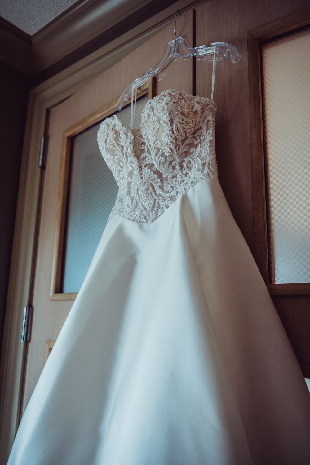 A white wedding dress with lace bodice and sweetheart neckline hangs elegantly against a wooden wall.
