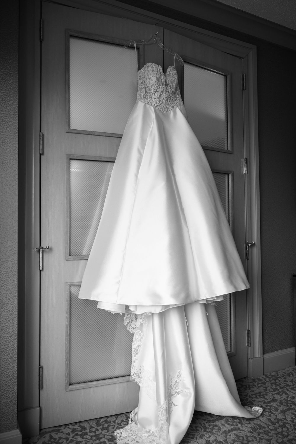 An elegant white wedding dress with lace detailing hangs on a door, photographed in black and white.