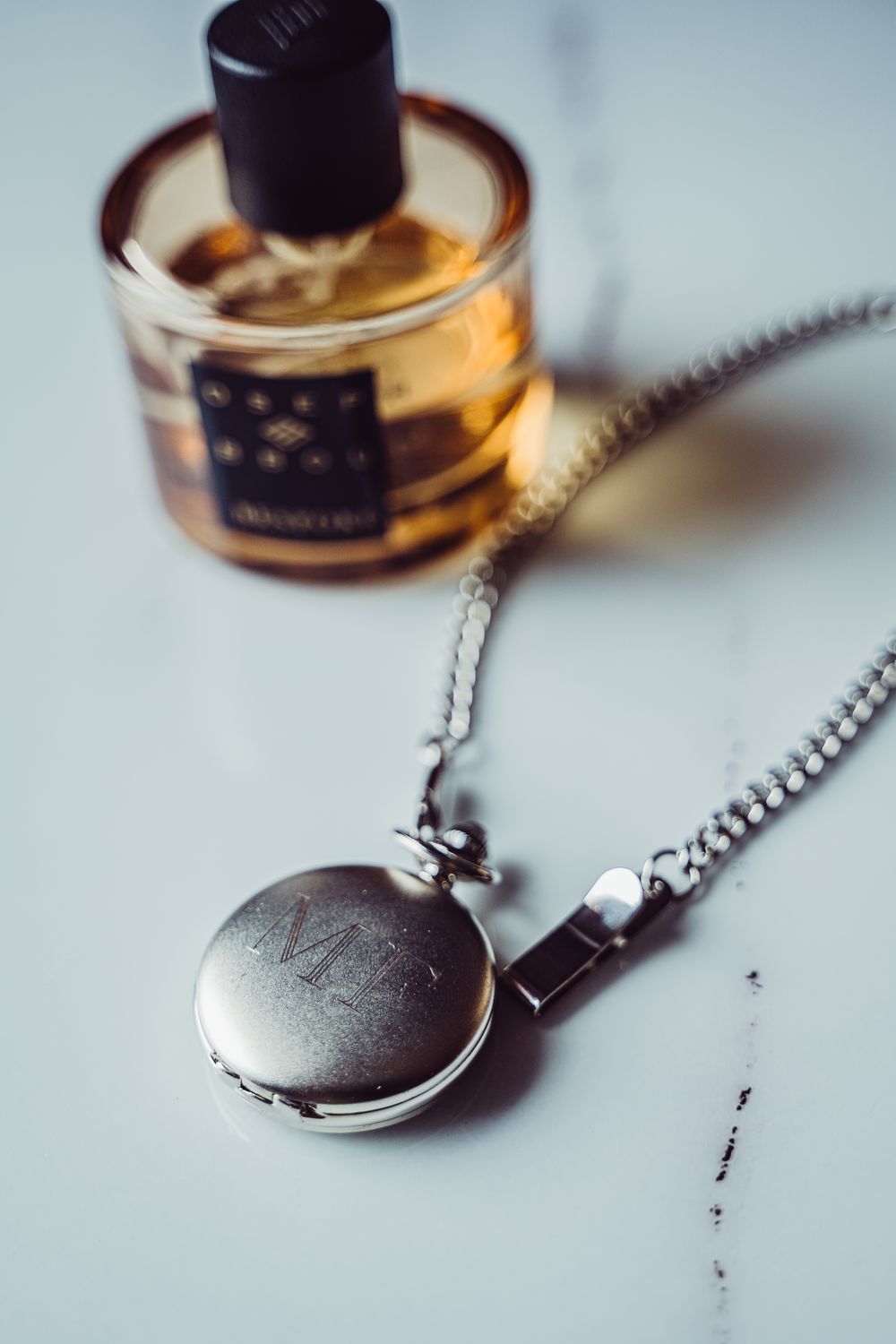 A vintage pocket watch on a silver chain sits beside a bottle of amber perfume or cologne on a light surface.