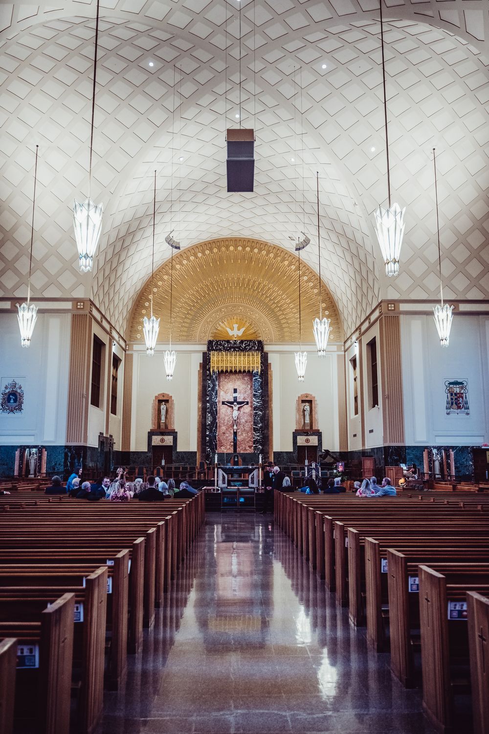 Panoramic view of a Catholic church interior with vaulted ceiling, hanging lights, and wooden pews leading to the altar.