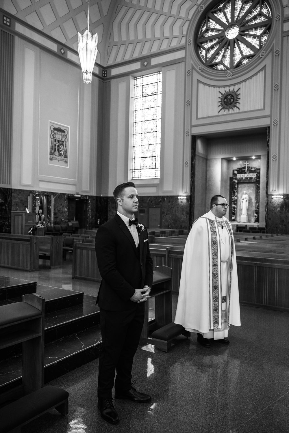 Black and white photo of a church interior with a groom and priest standing at the altar beneath a circular window.