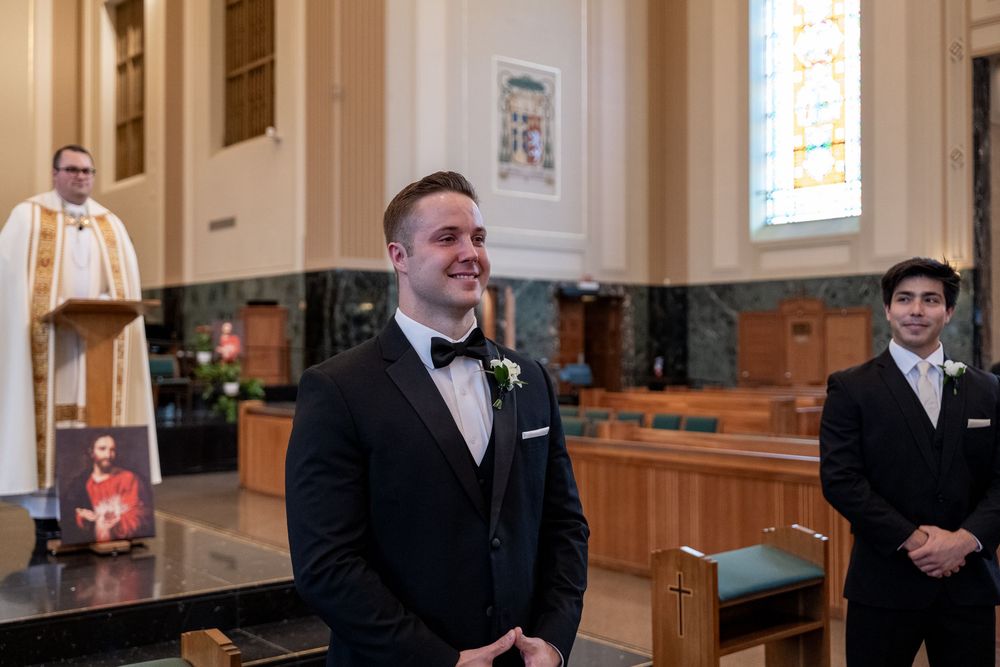 A groom stands at the altar in a church wearing a black tuxedo and bowtie during a wedding ceremony.