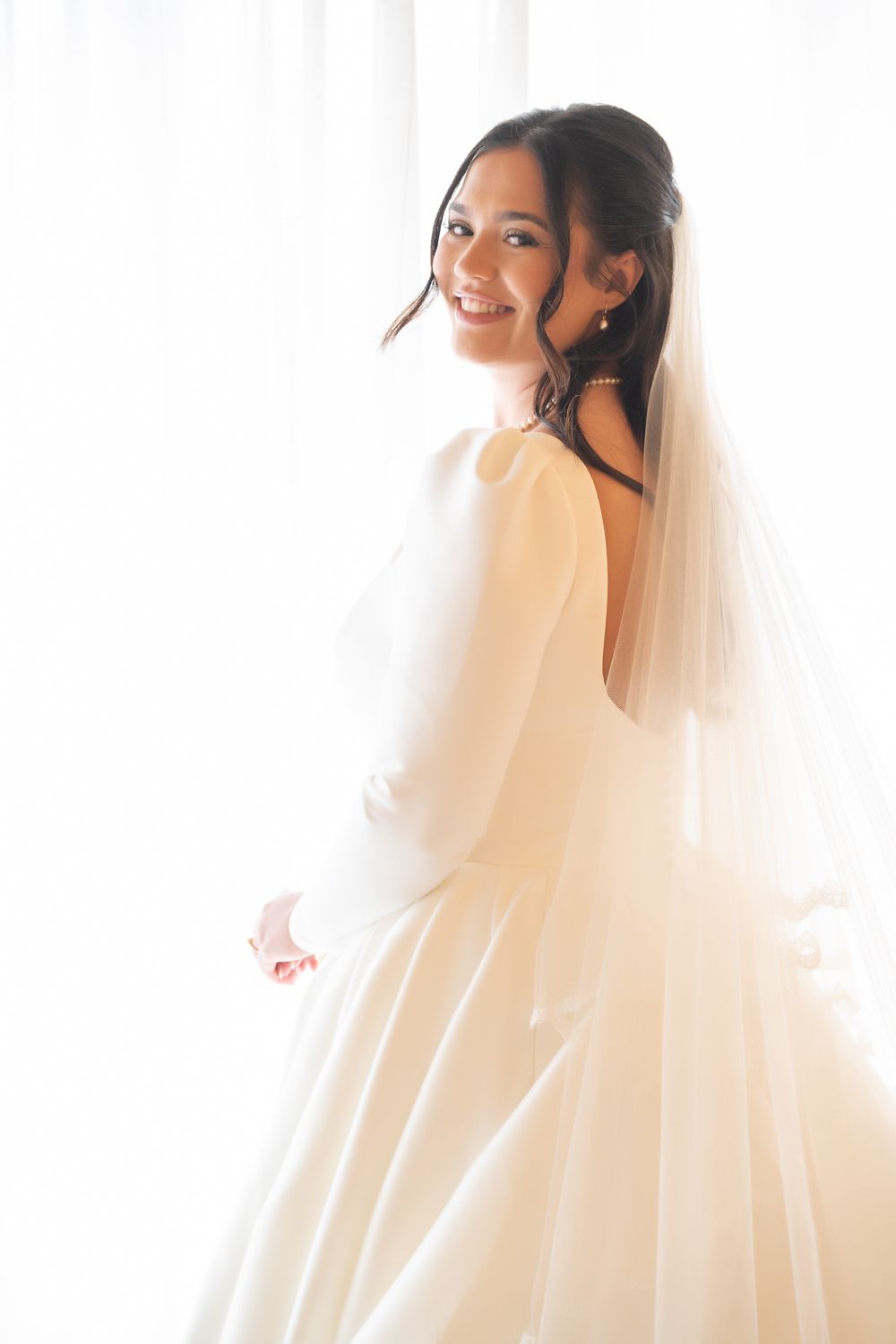 A bride in a flowing white wedding dress and veil poses elegantly in soft natural light.