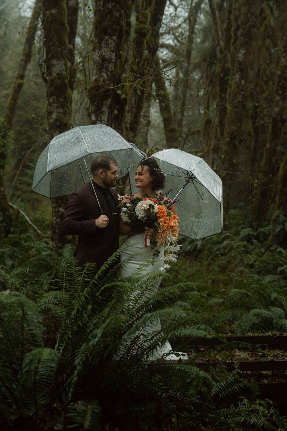 A romantic rainy day wedding photo shoot in a misty forest with clear umbrellas and orange bouquet.