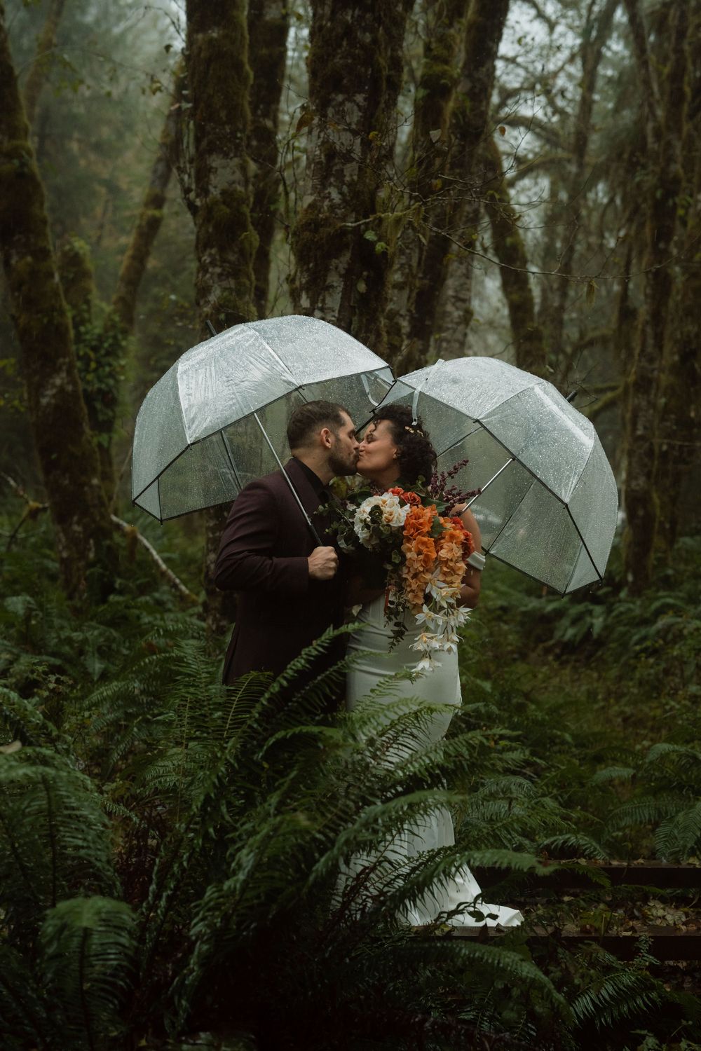 Newlyweds share an intimate moment under clear umbrellas in a lush forest setting.
