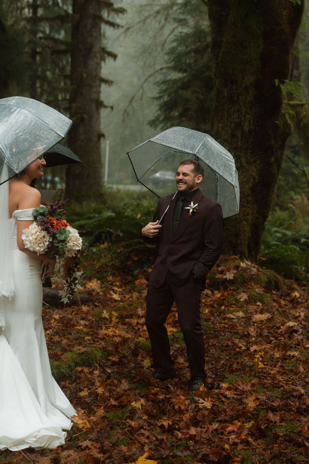 Wedding couple playfully poses with umbrellas among fallen autumn leaves in the forest.