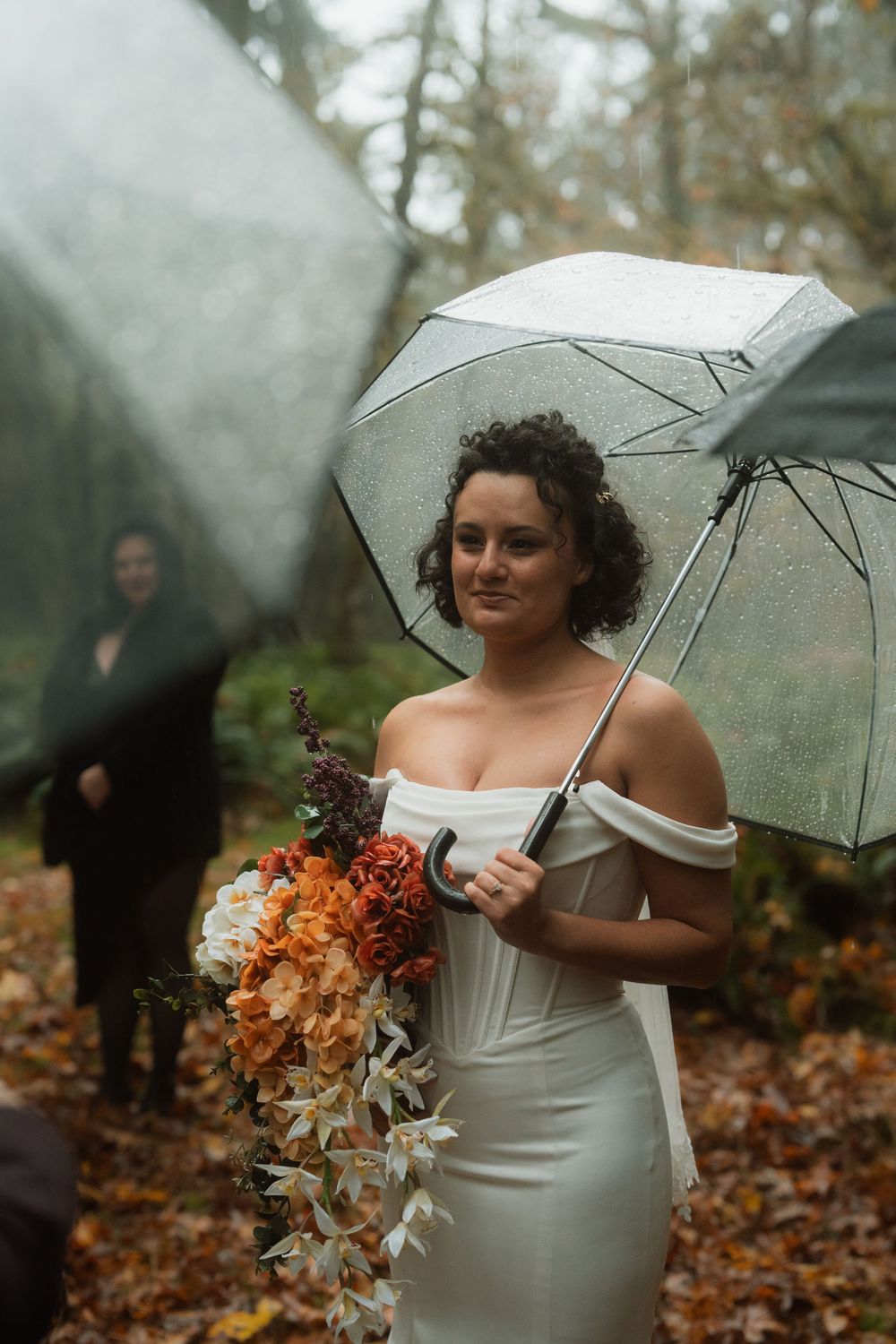 A bride holds a clear umbrella and autumn-colored bouquet while standing in a forest setting.