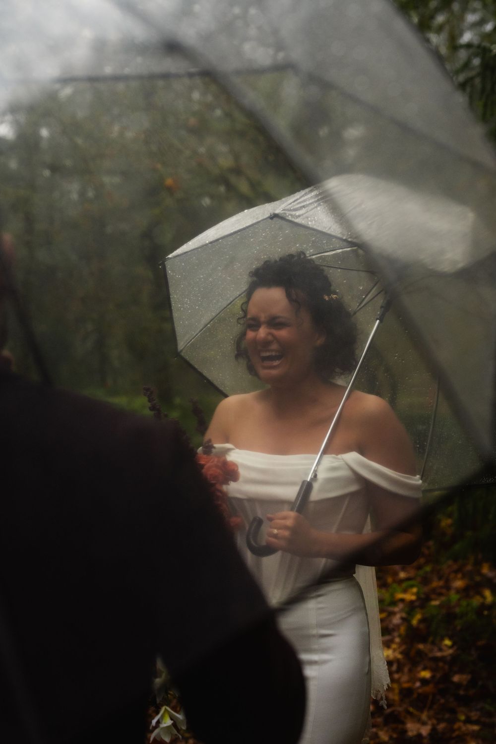 A person in a white dress smiles while holding a clear umbrella in a forest setting.
