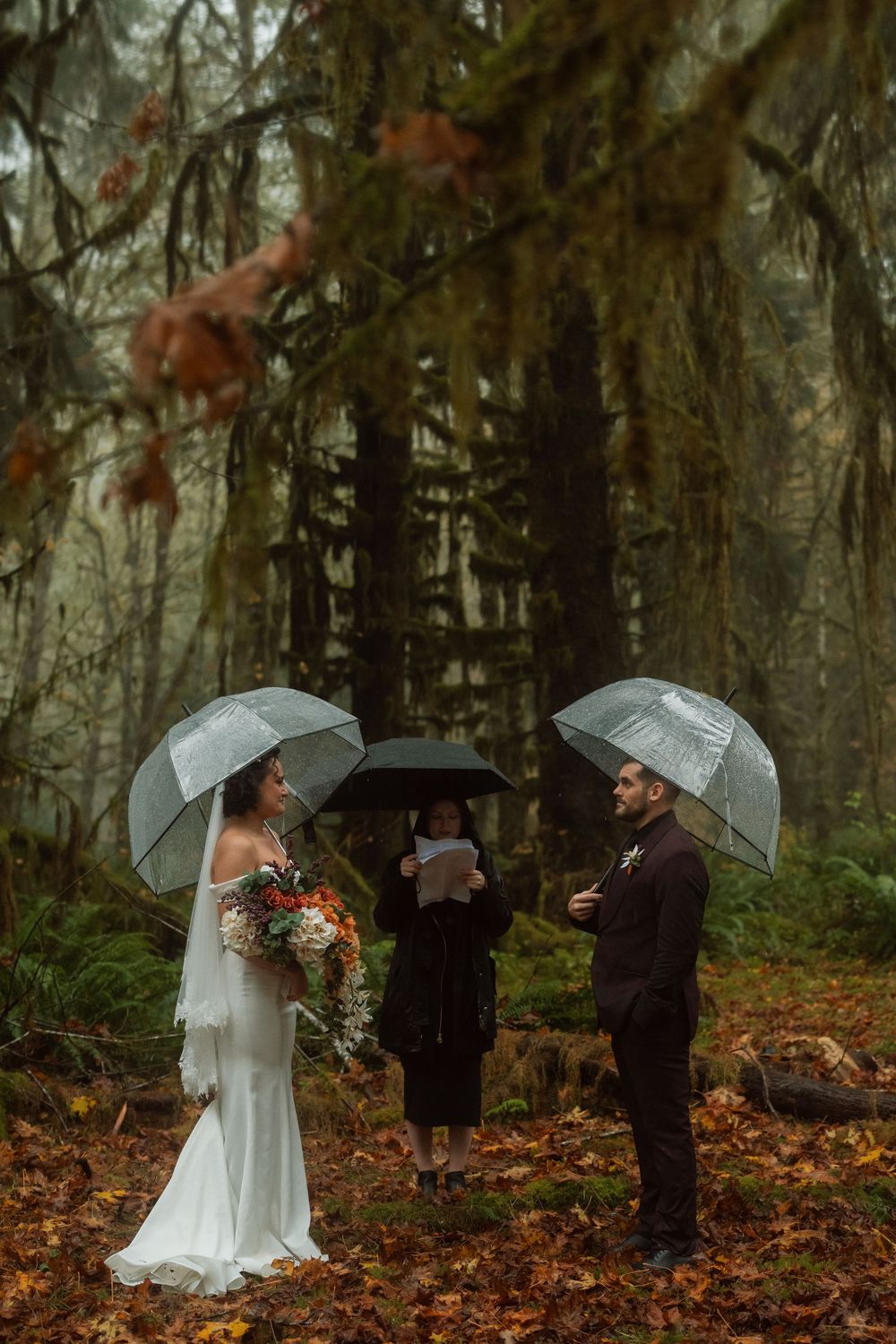 A wedding ceremony unfolds in a misty forest with guests holding clear umbrellas.