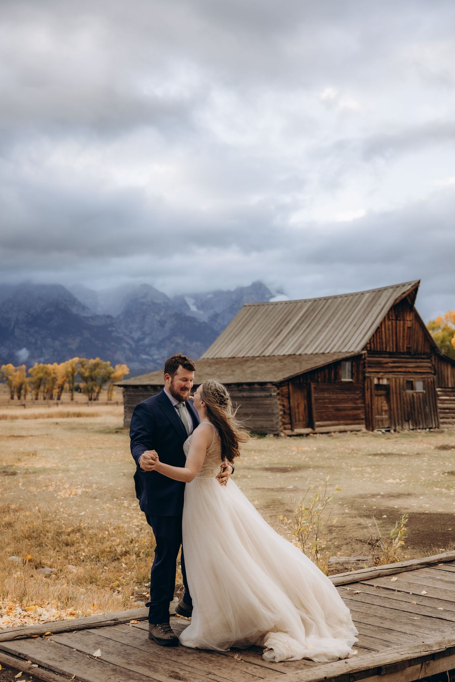 A couple dances on a wooden deck in front of a rustic barn with dramatic mountain backdrop and autumn colors.