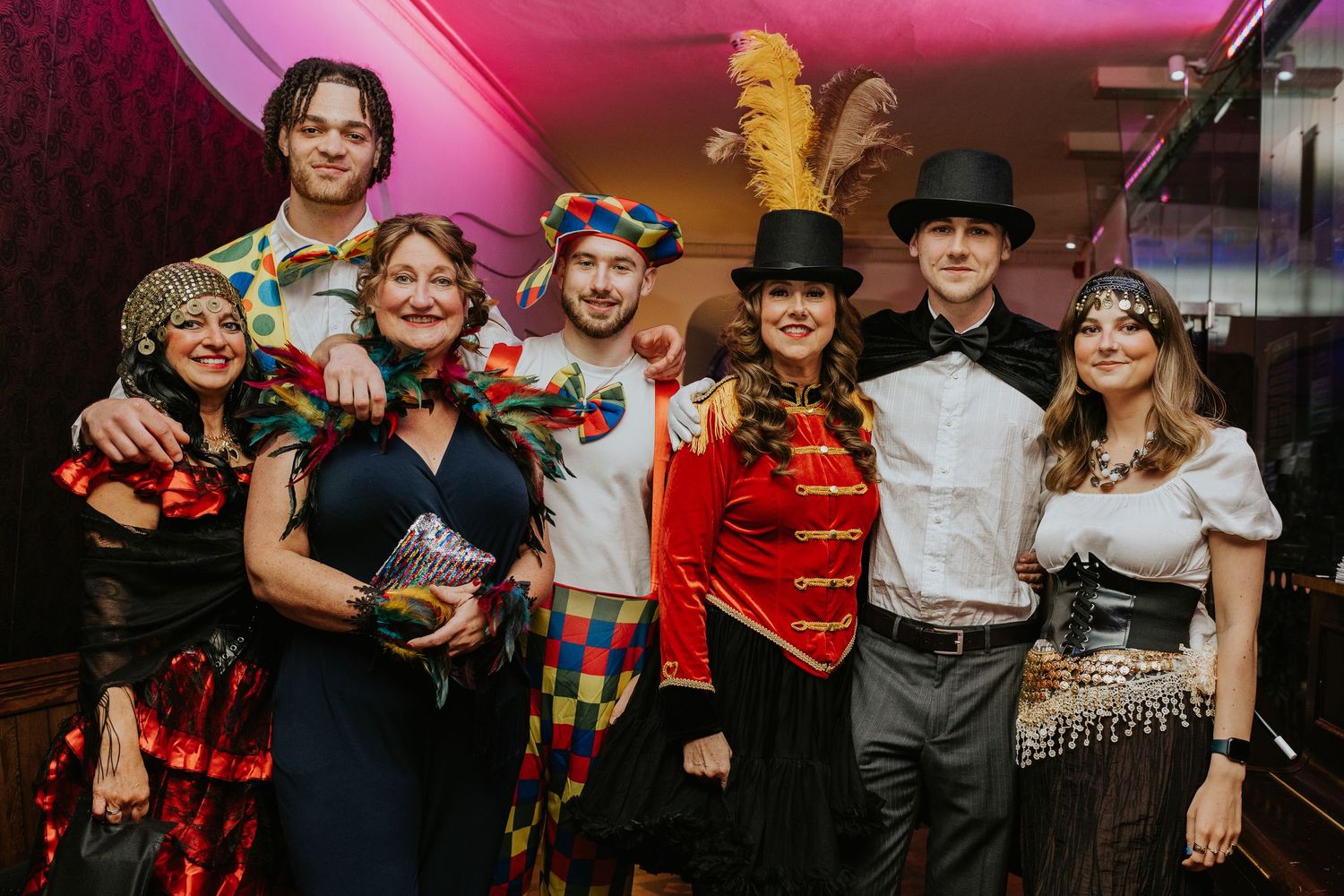 Group of people in festive costumes pose together at a party with pink lighting in the background.