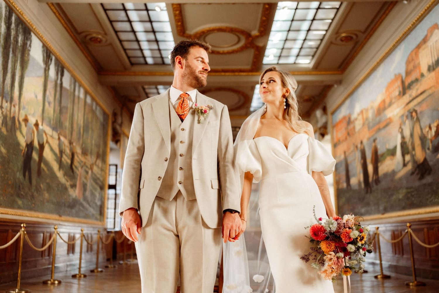 A couple stands in an elegant hallway with painted murals and skylights.