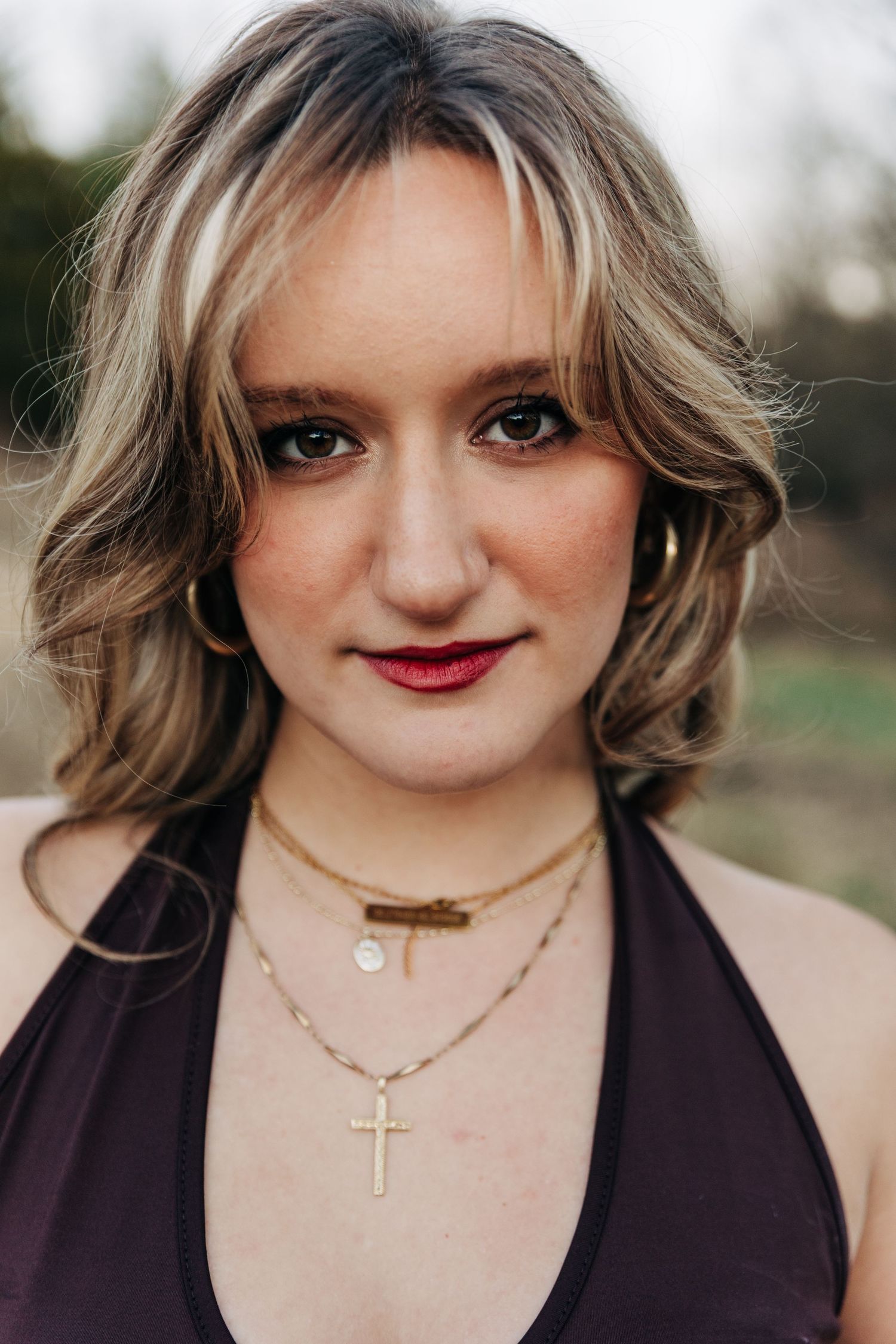 Close-up portrait of someone with wavy blonde hair wearing a brown top and layered gold necklaces, smiling outdoors.