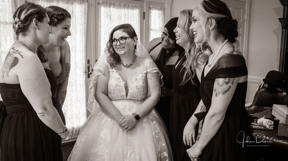 A candid black and white wedding moment shows a bride in glasses and white dress sharing laughter with attendants.