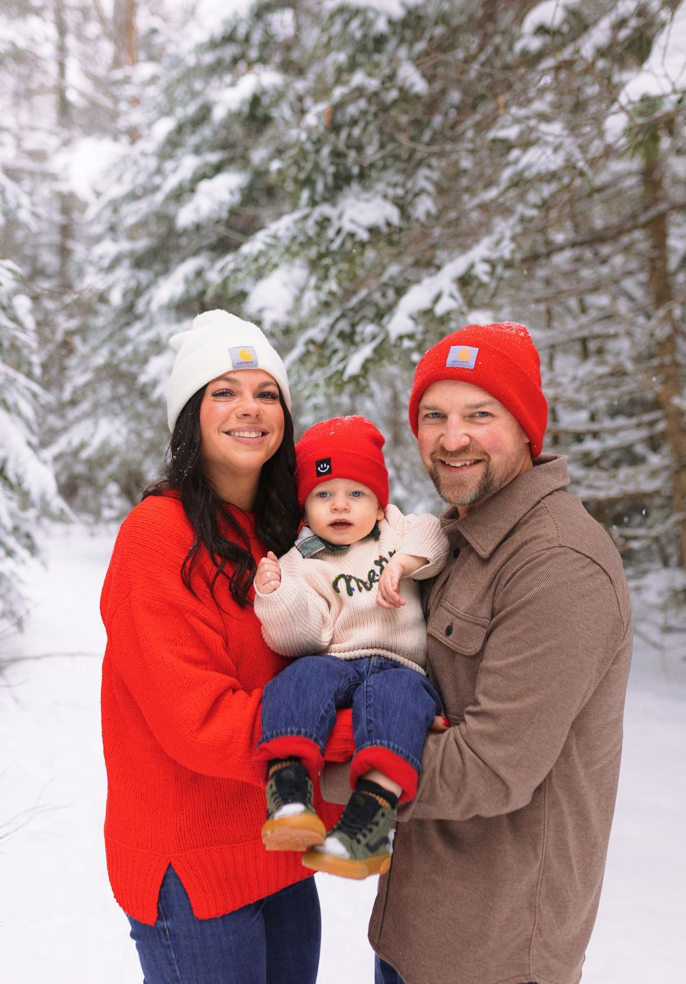 A family poses together in a snowy winter forest scene wearing red and brown winter clothing.