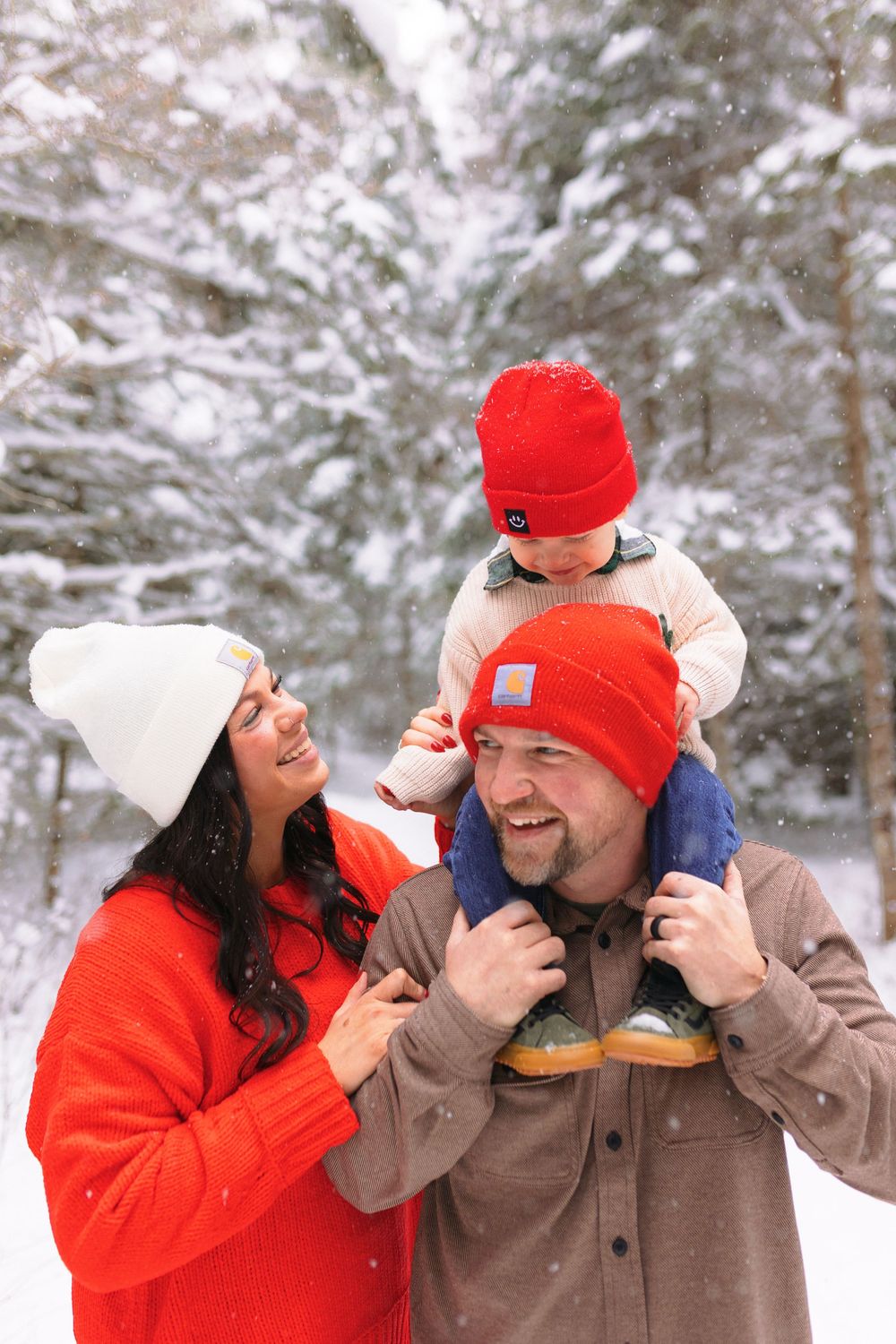 A family enjoys a winter day together in the snowy woods wearing matching red winter hats and warm coats.
