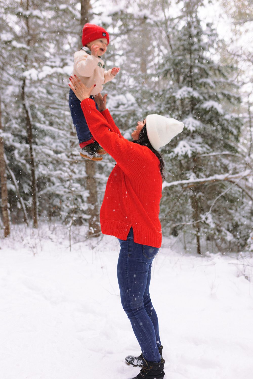 Person in bright red winter jacket and blue pants plays joyfully in deep snow against a backdrop of snow-covered evergreens.