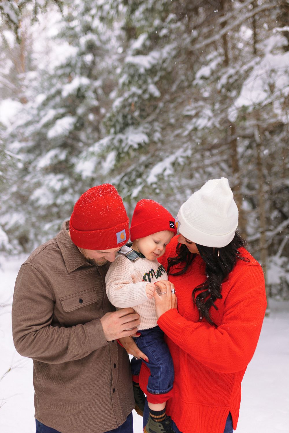 A family in winter attire shares tender moments in a snowy forest setting with red and brown clothing.