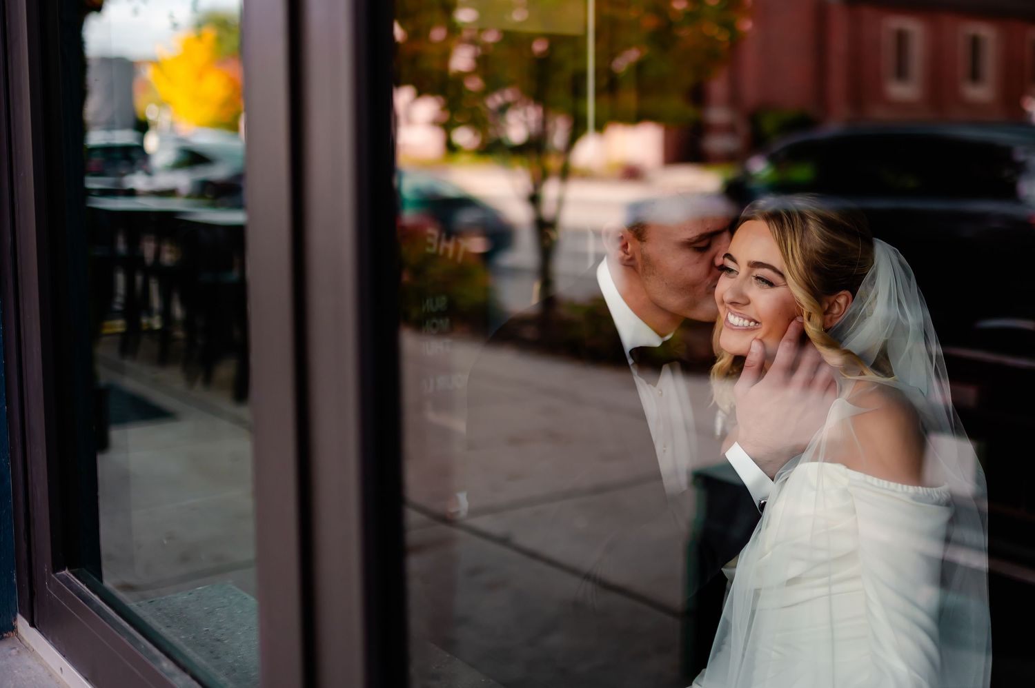 A bride and groom share a romantic moment against a window reflection in an urban setting.