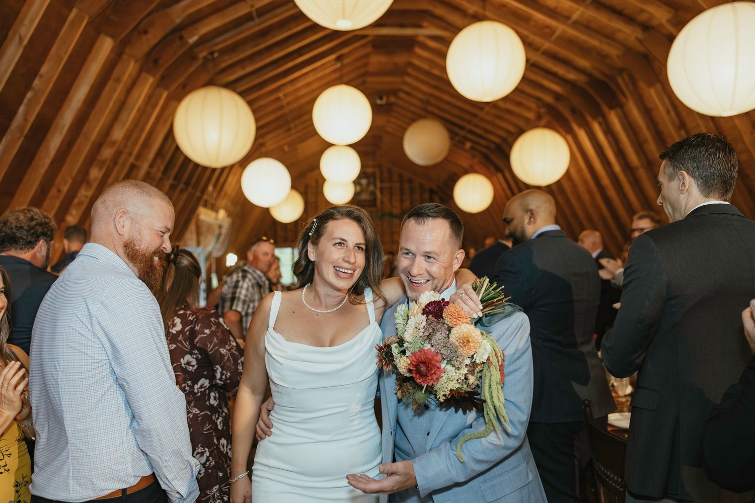 A rustic barn wedding reception with glowing paper lanterns illuminating the wooden beamed ceiling above guests.
