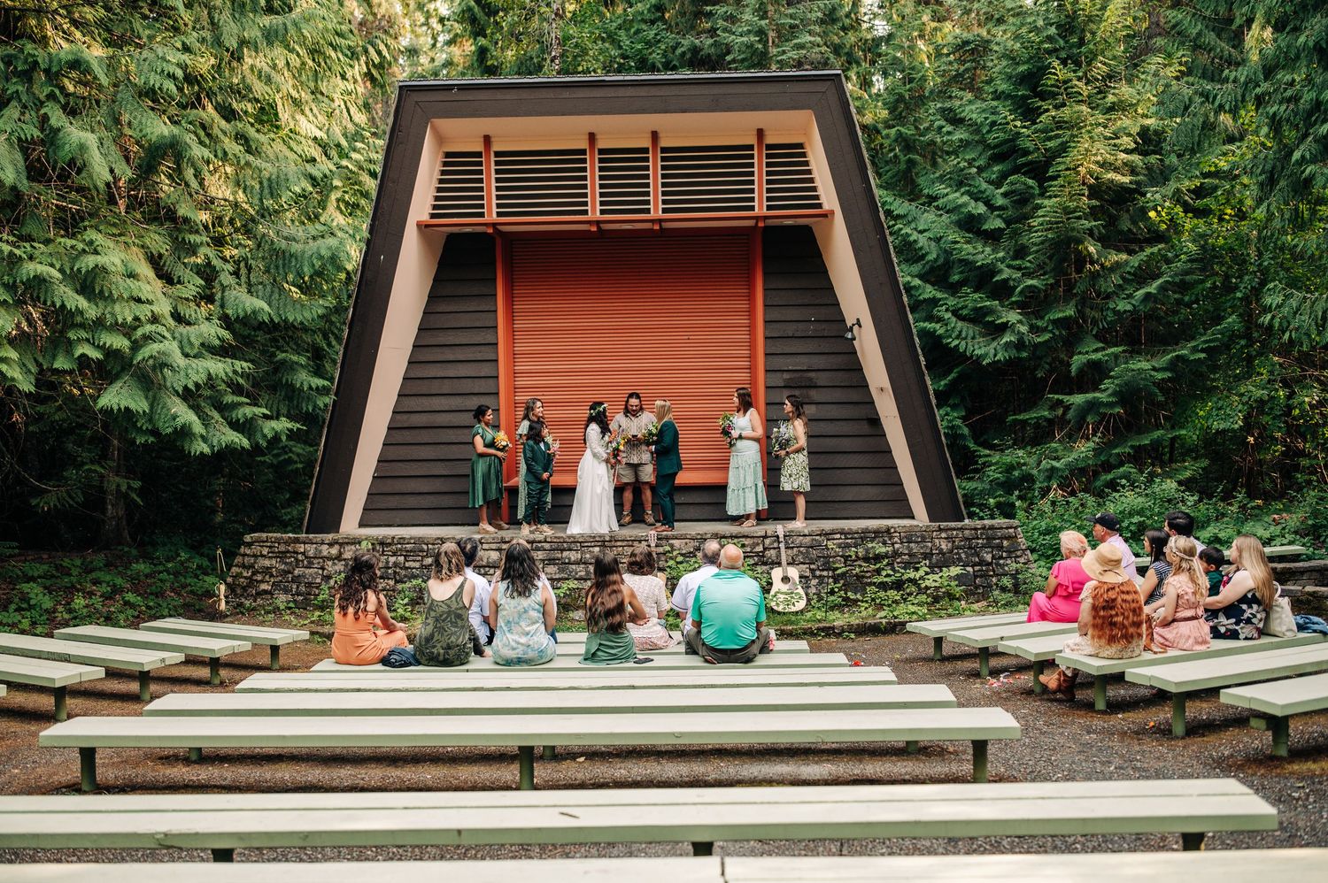 People sit on concrete steps in front of a rustic wooden amphitheater surrounded by lush green forest.