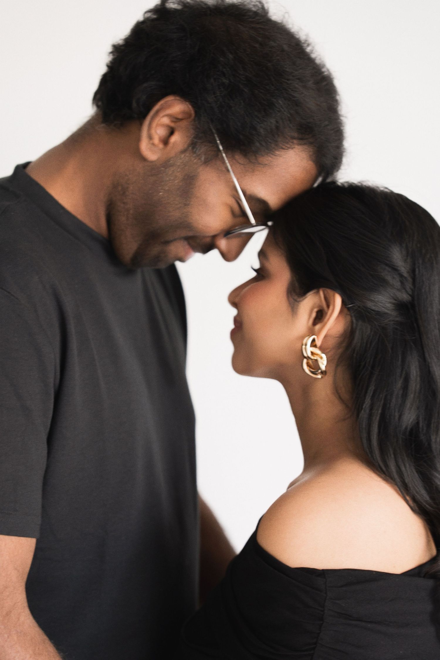 Intimate couple in black clothing share a tender forehead touch moment against white background.