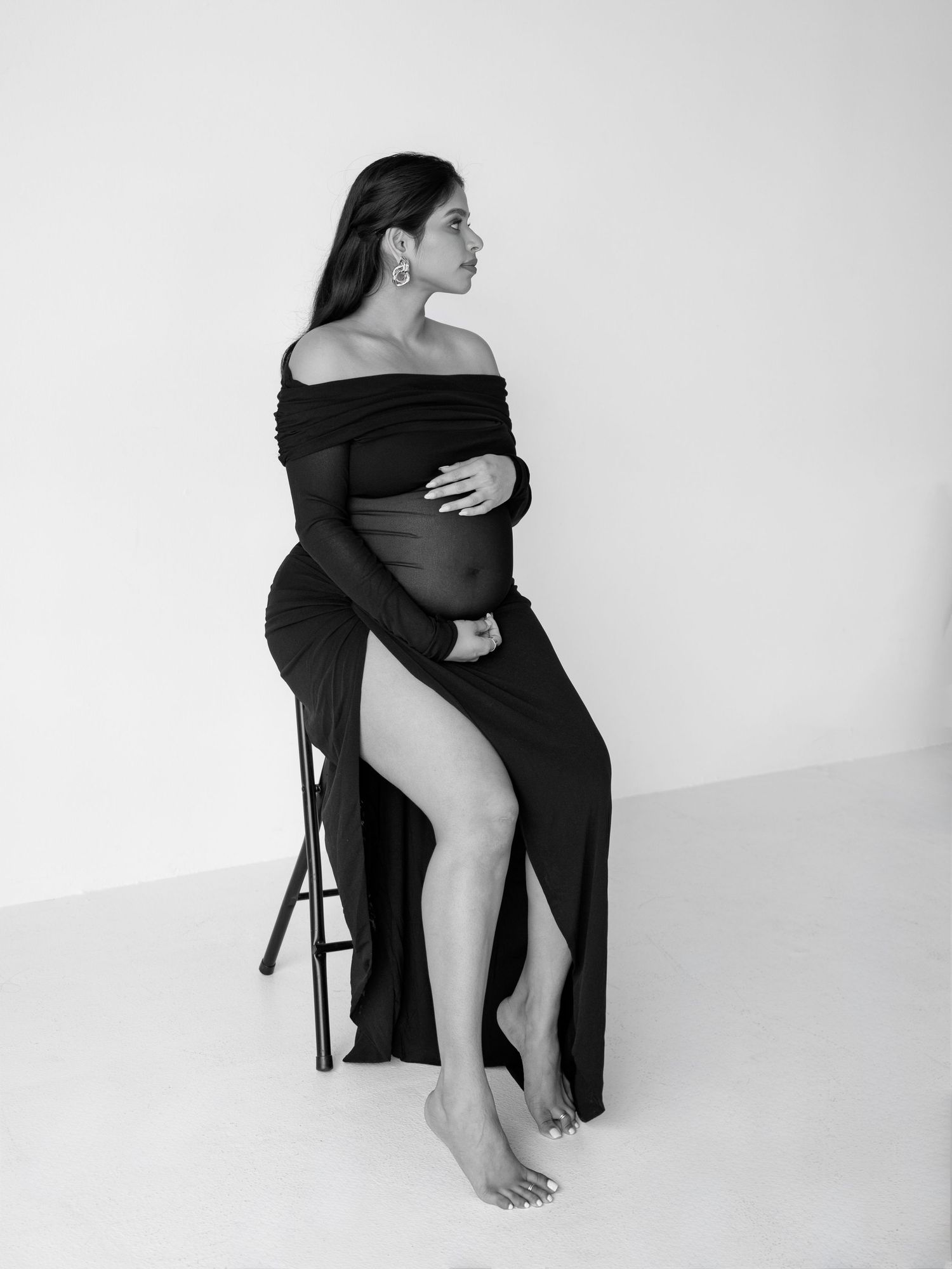 Black and white artistic maternity photo of a woman in an off-shoulder dress posing elegantly on a stool.