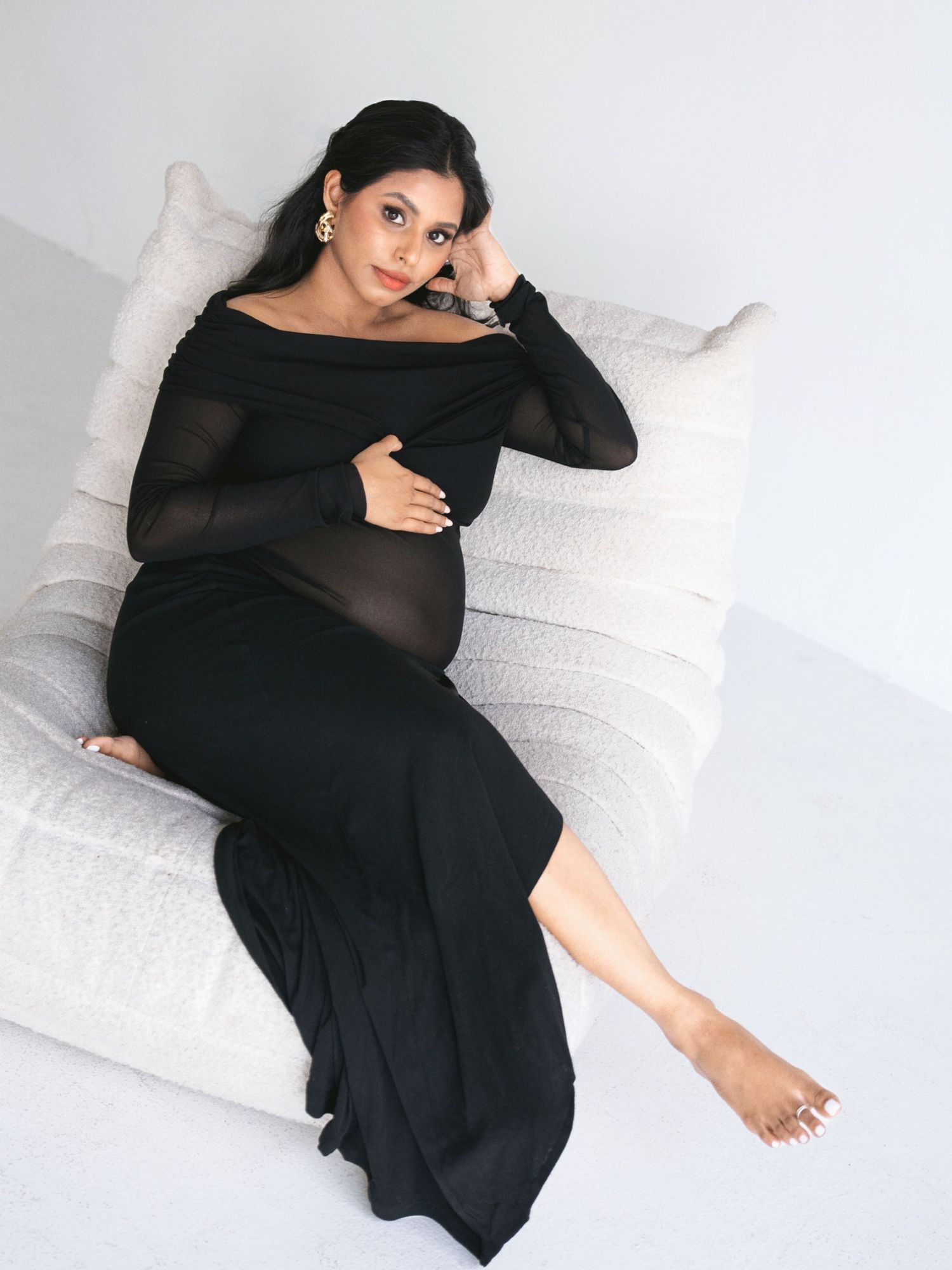 Expectant mother in elegant black dress reclines on white couch during maternity photoshoot.
