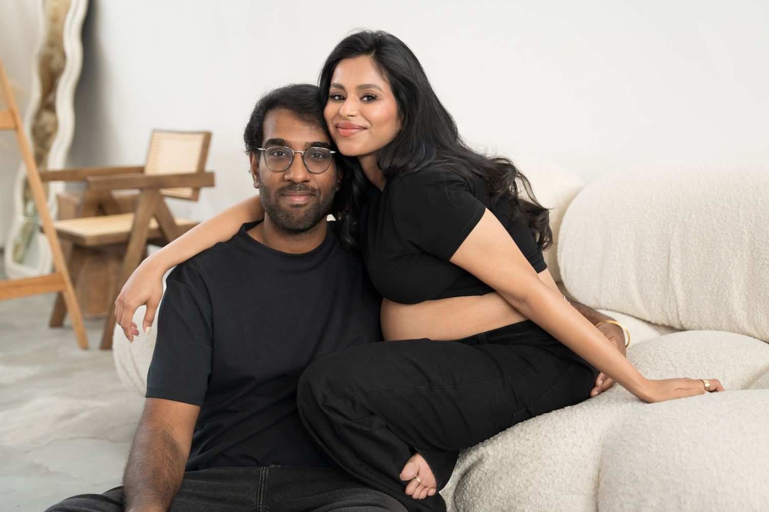 A couple in black clothing poses together on a couch in a modern living room setting.