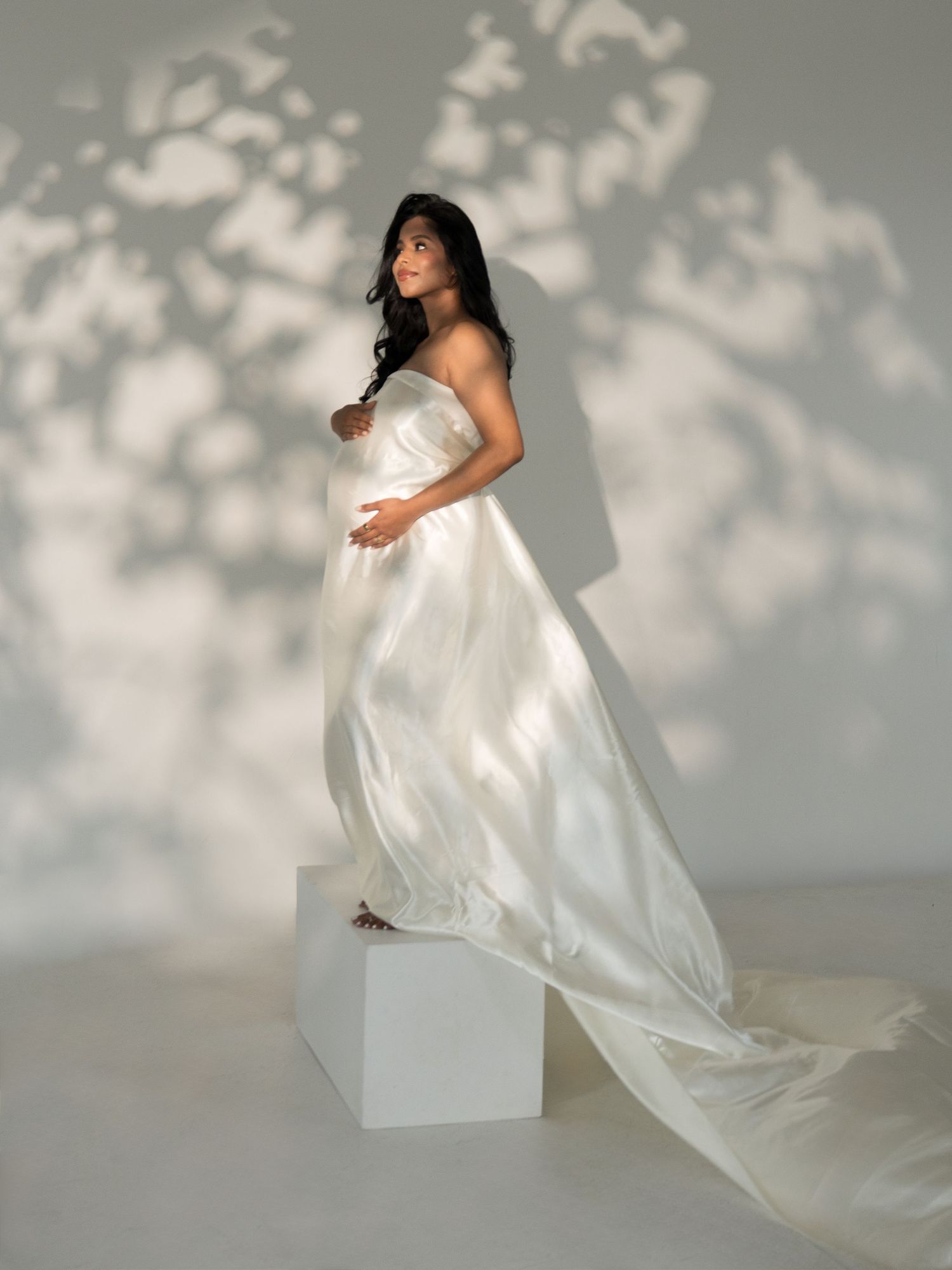 A maternity photoshoot featuring a flowing white dress against a soft grey backdrop with delicate shadow patterns.