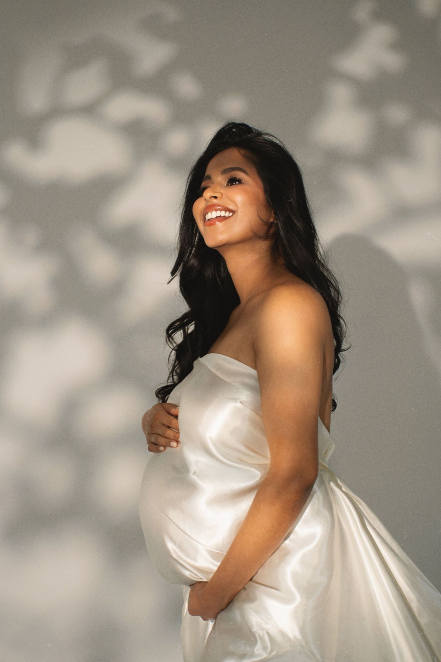 Expectant mother in flowing white dress smiles joyfully during maternity photoshoot with dappled lighting effects.