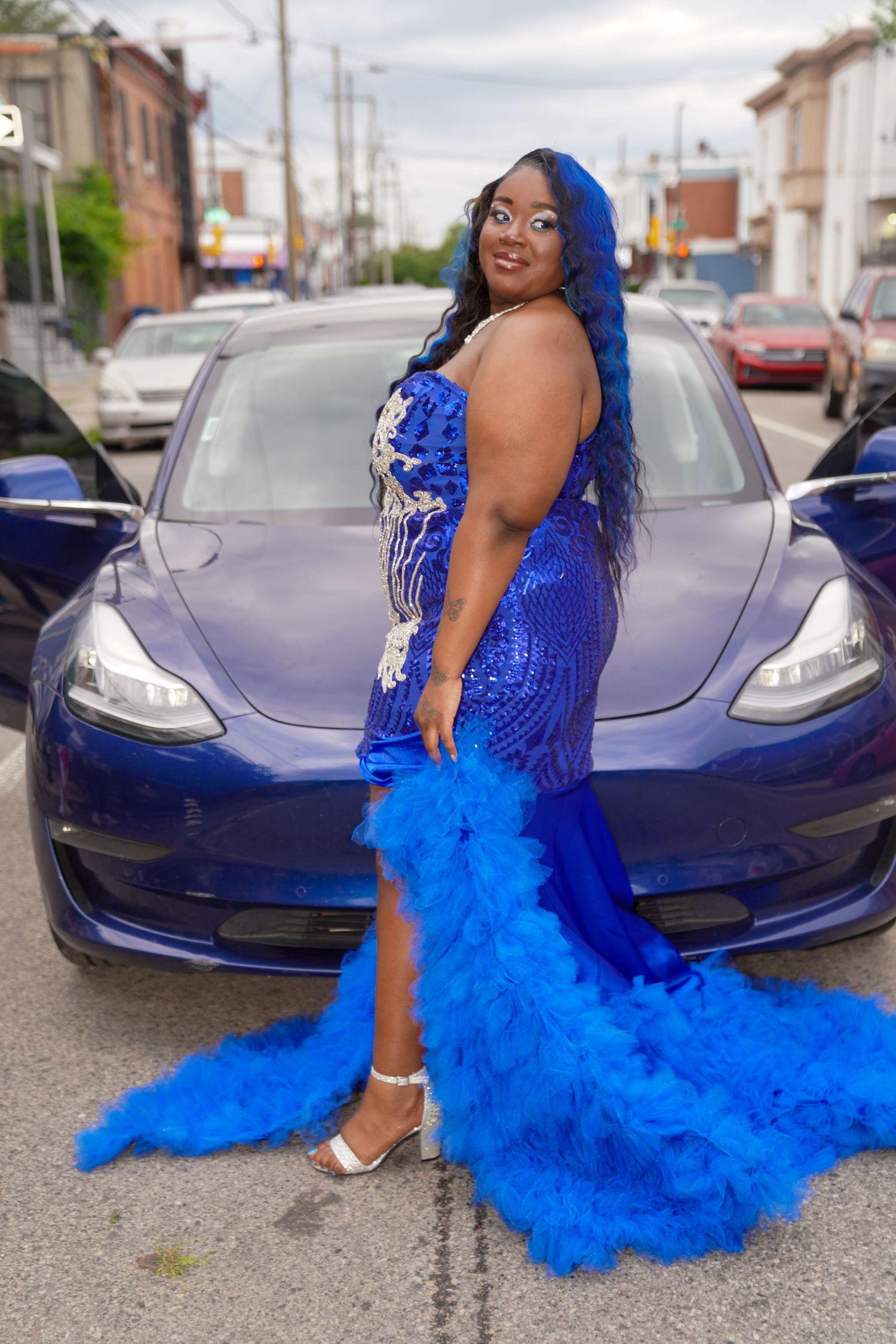 A vibrant royal blue tulle gown with lace bodice photographed against a Tesla Model 3 car on a city street.