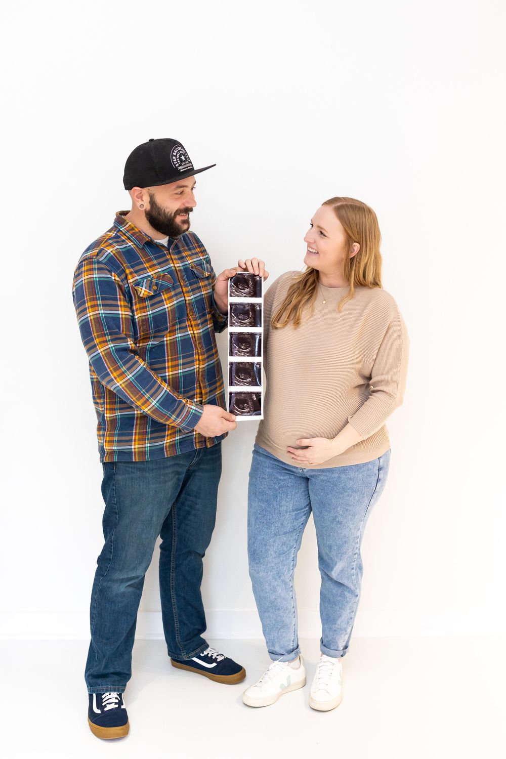 A couple reviews ultrasound images together while standing against a white wall in casual attire.