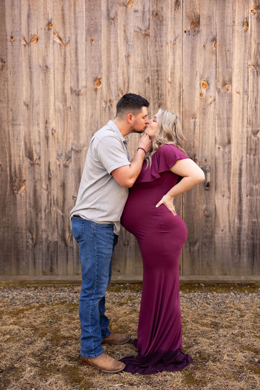 A couple shares a tender moment against a rustic wooden fence backdrop, with the woman wearing a burgundy maternity dress.
