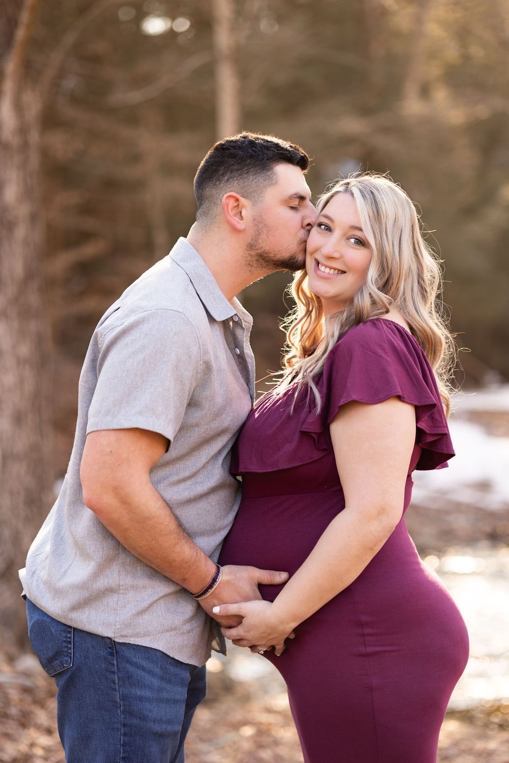 A couple shares a tender moment outdoors as they celebrate their pregnancy in a burgundy dress and casual attire.