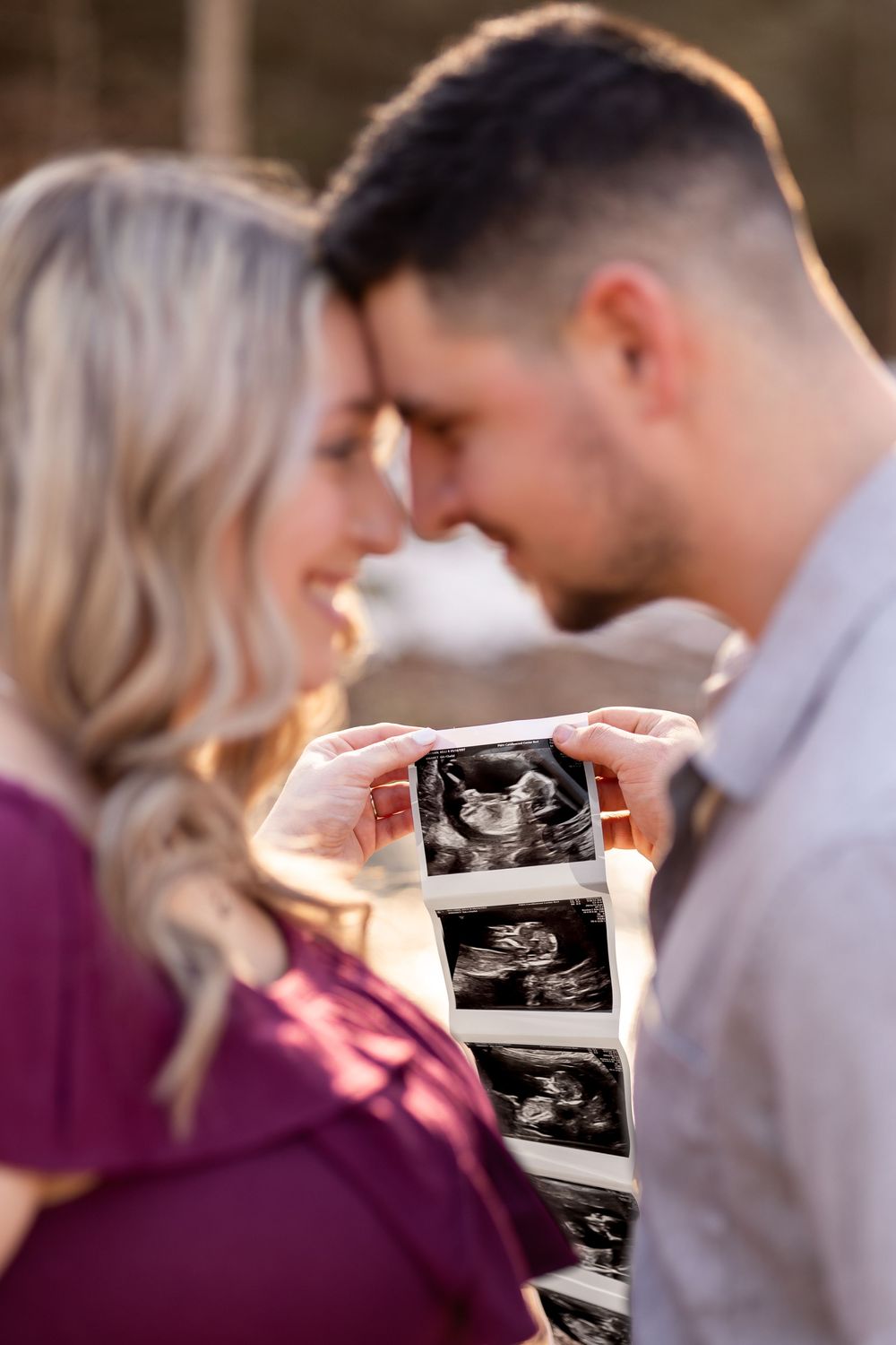 A couple shares an intimate moment while holding ultrasound photos during a pregnancy announcement photoshoot at sunset.