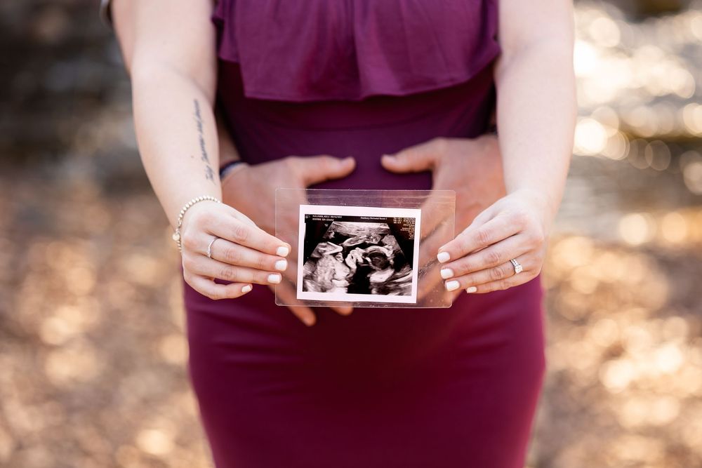 Hands holding an ultrasound photo against a burgundy dress in outdoor natural lighting.