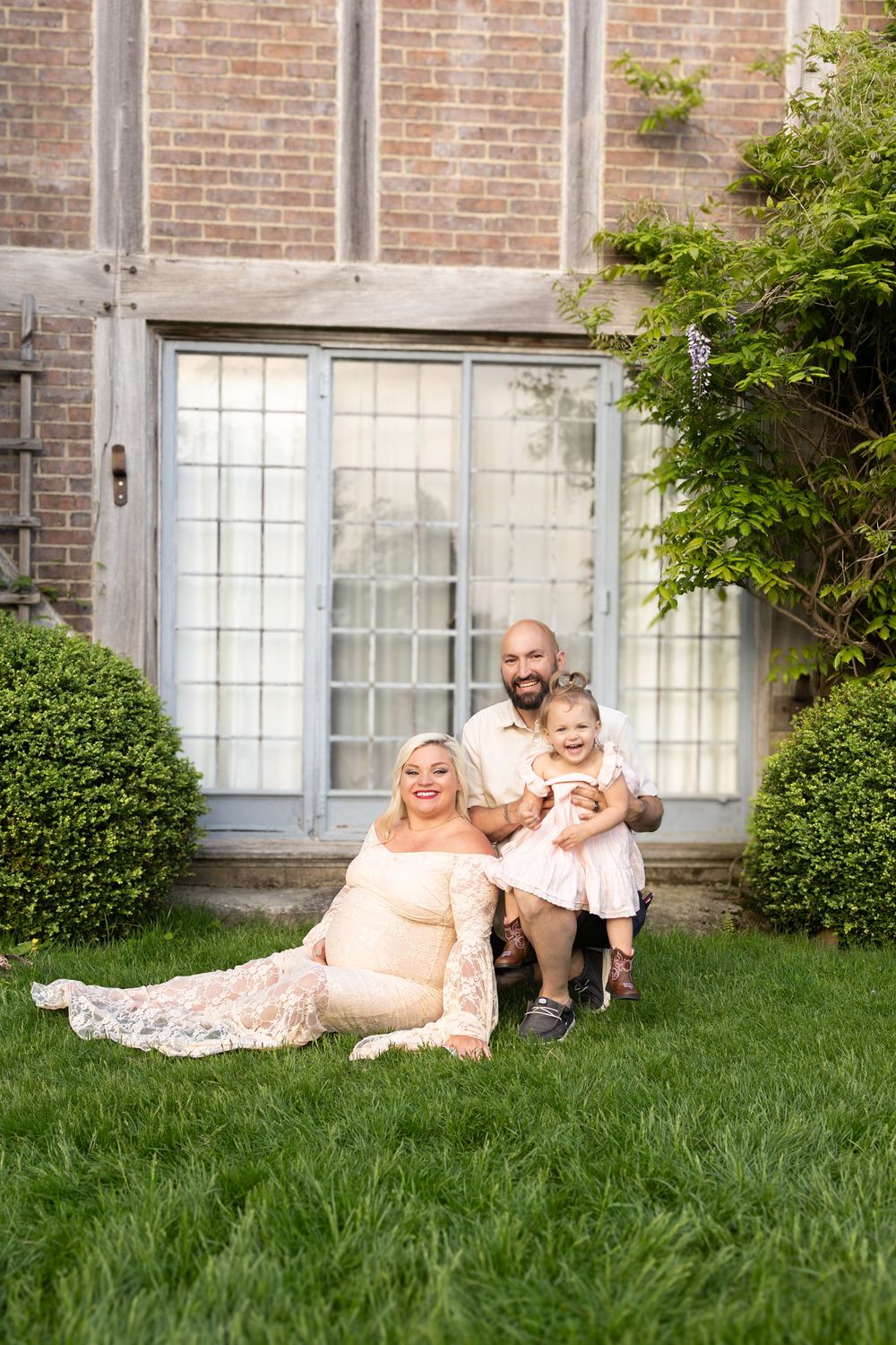 Family relaxes together on green lawn outside brick building with white windows on sunny day.