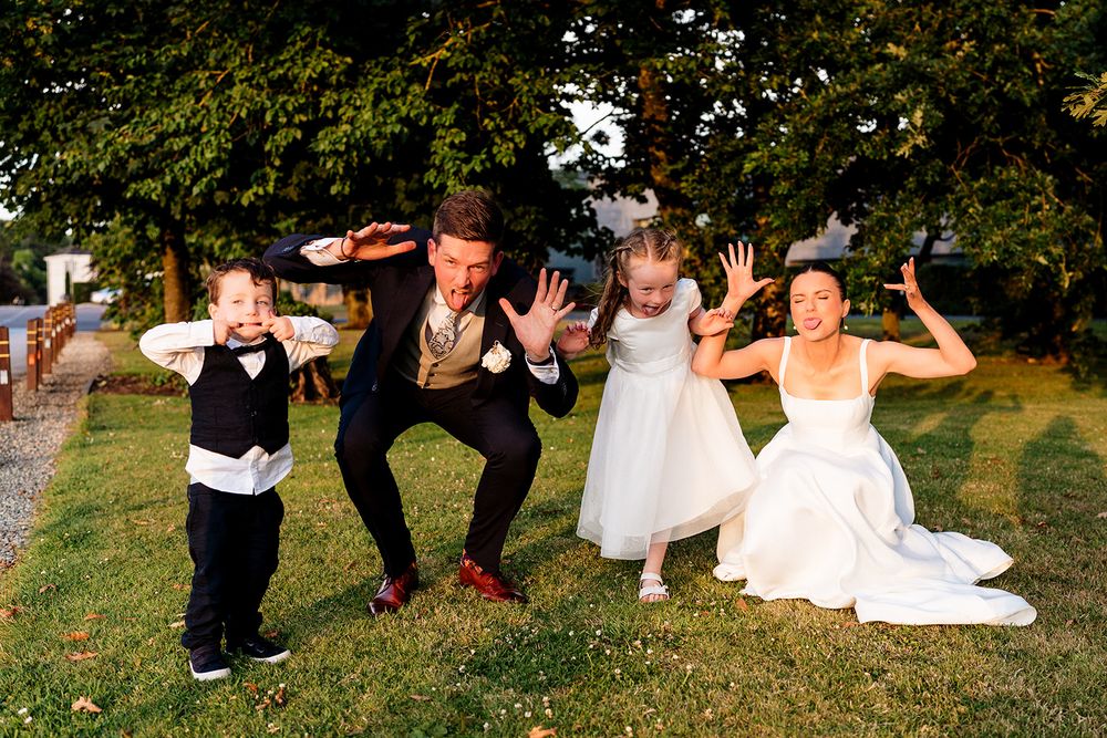 A fun wedding party poses playfully on the grass with their hands raised during an outdoor celebration.