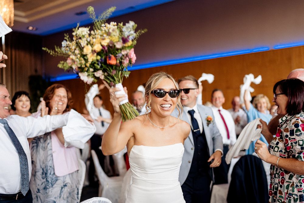 A dance floor full of wedding guests dancing at a reception with floral decorations and blue uplighting overhead.