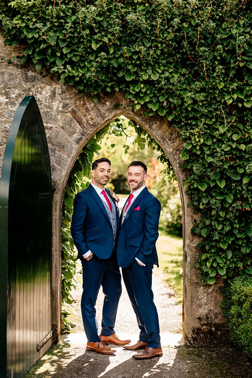 Two people in navy suits pose together in a romantic garden archway setting with ivy covered stone walls.