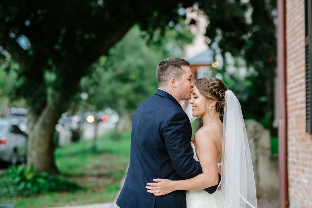A bride and groom share an intimate moment outdoors near a brick building and trees.