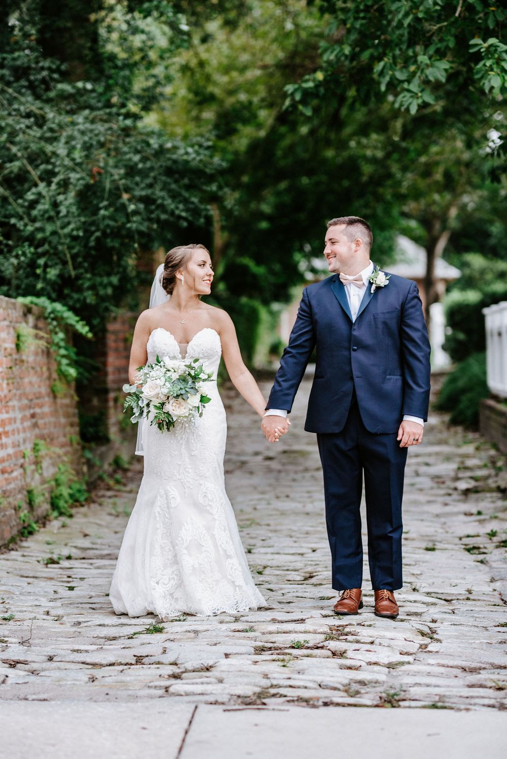 A bride and groom hold hands while walking along a cobblestone path surrounded by lush greenery.