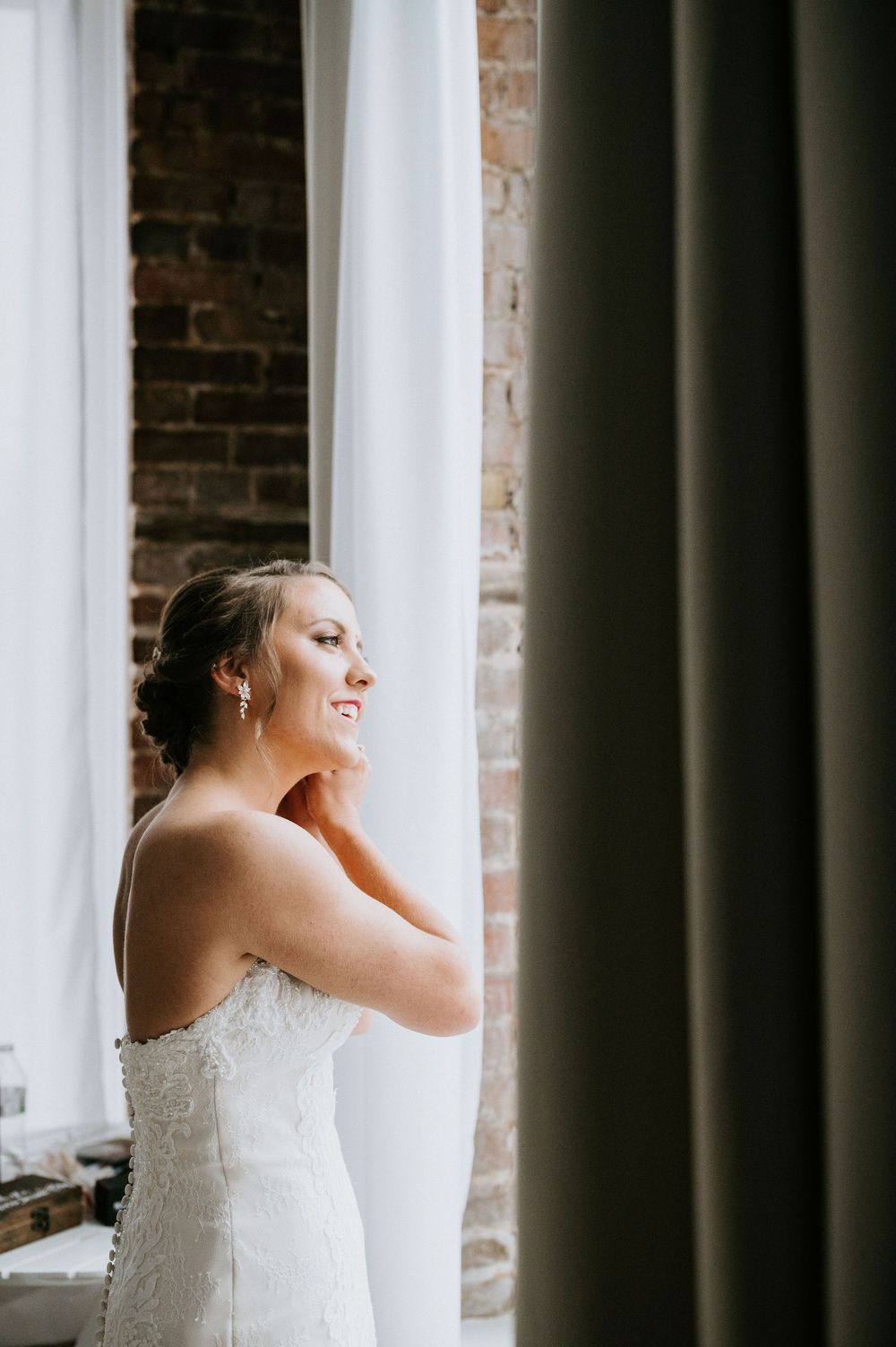 Bride in elegant white strapless gown poses by window with natural light streaming in against exposed brick wall.