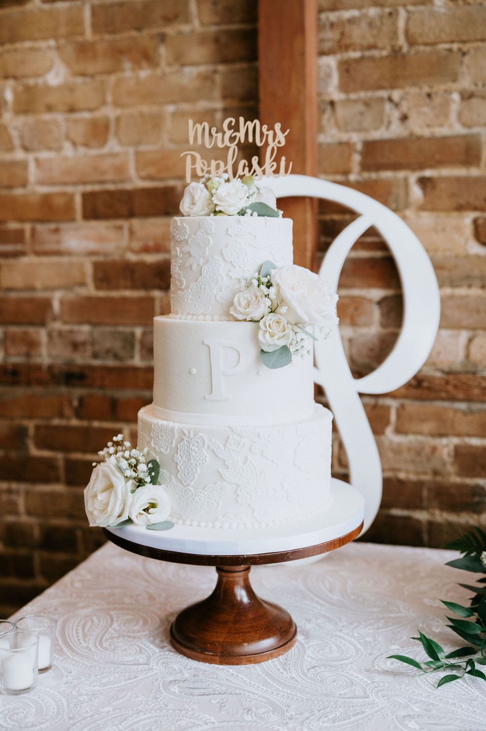 Elegant white three-tiered wedding cake with lace details and floral accents displayed on wooden stand against rustic brick wall.
