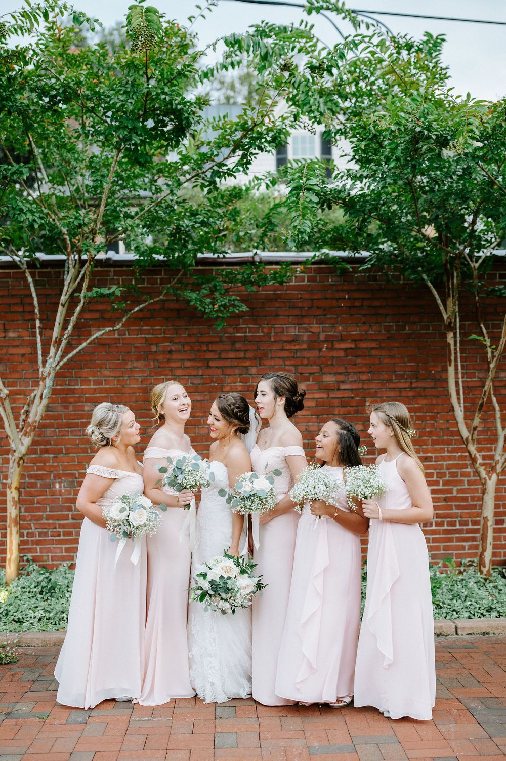 A wedding party in blush pink dresses poses together against a brick wall with green foliage overhead.