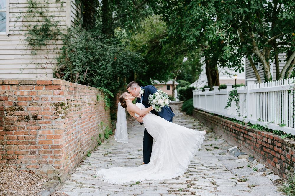 A romantic couple shares a kiss on a cobblestone path beside a historic brick wall and white picket fence.
